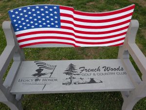 EE3F8702-10CC-4C82-830C-93334F822CC2 | 3i Graphics & Signs Grey bench with a large American flag motif on the backrest and an engraved plaque on the seat reading 'Legacy of Honor' at French Woods Golf & Country Club, on grass.